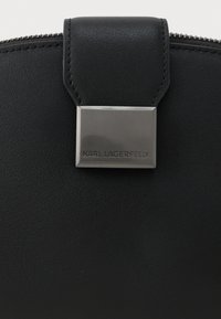 Melnas ādas somas detaļa ar sudraba metāla sprādzi, uz kuras ir ierakstīts "Karl Lagerfeld", un redzama rāvējslēdzēja augšējā mala.