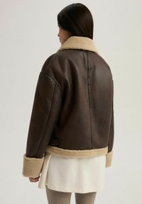Brązowa skórzana kurtka z beżowym kołnierzem i mankietami z owczej wełny. Projekt charakteryzuje się pudełkowym krojem oraz widocznym szwem wzdłuż szwów.