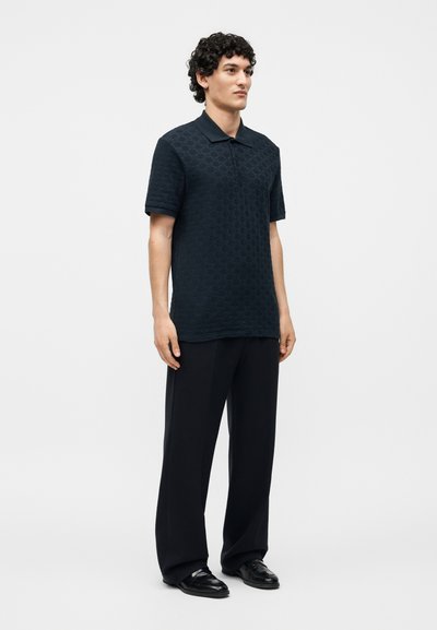 Άνδρας στέκεται φορώντας ένα σκούρο υφασμάτινο κοντομάνικο πόλο, χαλαρό μαύρο παντελόνι και μαύρα παπούτσια μπροστά σε απλό φόντο.