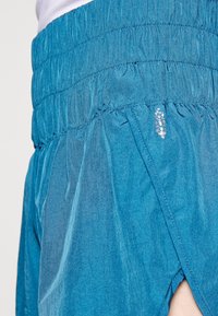 Blå atletiska shorts med samlad elastisk midja, med en skimrande yta och en sidofåra för ventilation. Lätt, andningsbart tyg.