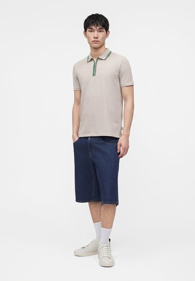 Jeune homme debout portant un polo beige à manches courtes avec bordure verte, un short en jean foncé jusqu’aux genoux, des chaussettes blanches et des baskets blanches.