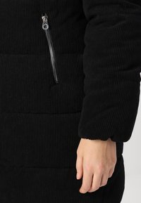 Veste en velours côtelé noir avec une poche latérale zippée, une texture côtelée et un design rembourré, avec une main visible reposant sur le tissu.