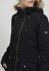 Black outerwear jacket with a faux fur-lined hood, contrasting brown trim, multiple pockets, and snap/button closures. Textured fabric.
