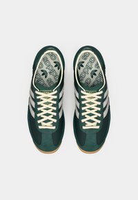 Dark green sneakers with light grey stripes, textured mesh upper, and cream laces. Rubber outsole with branding on the tongue and insole.