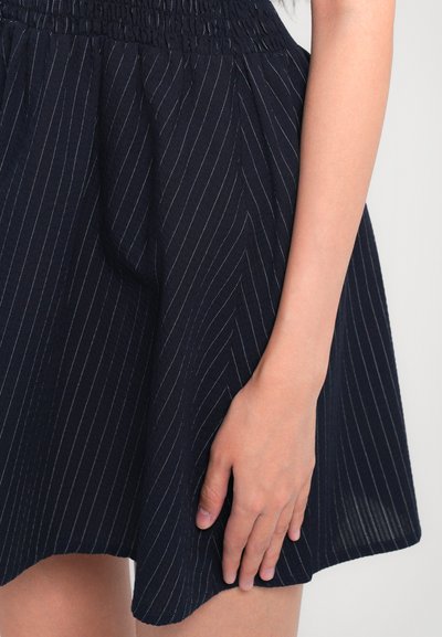 Tengerészkék ruha texturált felülettel, függőleges fehér csíkokkal és elastikus derékkal. Kéz finoman pihen a anyagon.