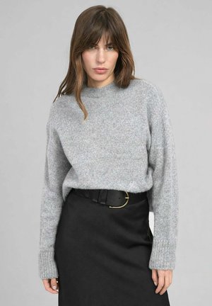 Femme aux cheveux bruns mi-longs portant un pull en maille gris rentré dans une jupe noire avec une ceinture noire.