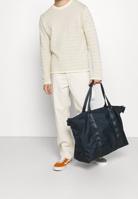 Svart nylon toteväska med tjocka, släta handtag och siddragkedjor, i kombination med en krämfärgad, texturerad tröja och ljusa byxor.