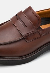 Zapato de piel marrón con un acabado suave, que cuenta con un diseño tipo slip-on, detalles de costura y paneles laterales elásticos, montado sobre una suela de goma negra.