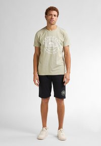 Camiseta de algodón verde claro con un diseño gráfico blanco, combinada con pantalones cortos negros que presentan un logo circular. Zapatillas blancas completan el conjunto.