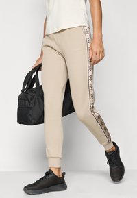 Άτομο που φοράει μπεζ παντελόνια με διακοσμητικές ρίγες στο πλάι, μαύρα αθλητικά παπούτσια, κρατώντας μια μαύρη τσάντα σωματοφύλακα, περπατώντας μπροστά σε μια απλή φόντο.