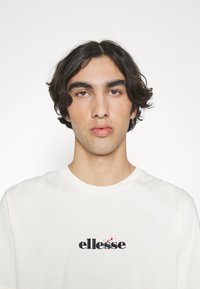 Weißes T-Shirt mit kurzen Ärmeln, das ein schwarzes, gesticktes "ellesse"-Logo mit floralen Akzenten auf der Brust zeigt. Glatte Baumwollqualität.