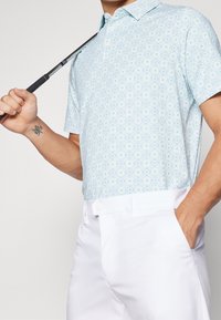 Světle modré polo tričko s geometrickým vzorem v zelené a navy. Krátké rukávy, s límečkem, spárované s bílými kalhotami. Golfová hůl držena v ruce.