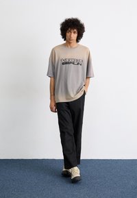 Light brown to grey gradient oversized t-shirt with graphic text, paired with black trousers and beige hiking boots on a blue carpet.