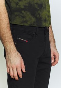 Jeans in denim neri con un design aderente, caratterizzati da un'etichetta rossa sulla tasca frontale e una camicia tie-dye verde a trama parzialmente visibile.