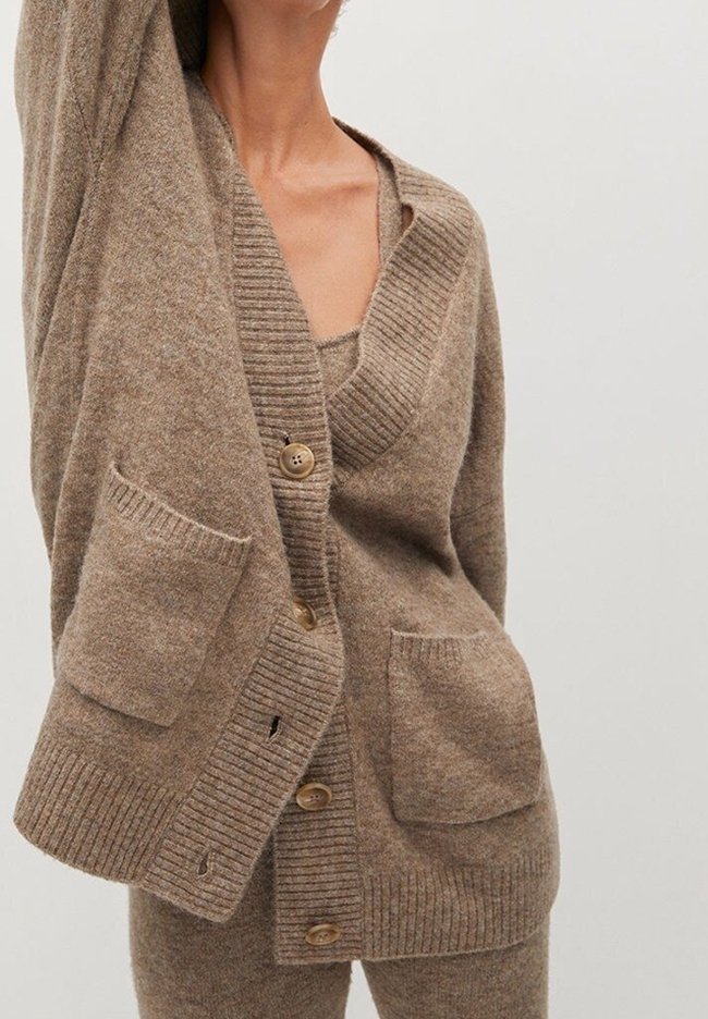 Femme portant un cardigan marron doux avec de gros boutons et des poches devant, par-dessus un haut et un pantalon assortis, sur un fond uni.