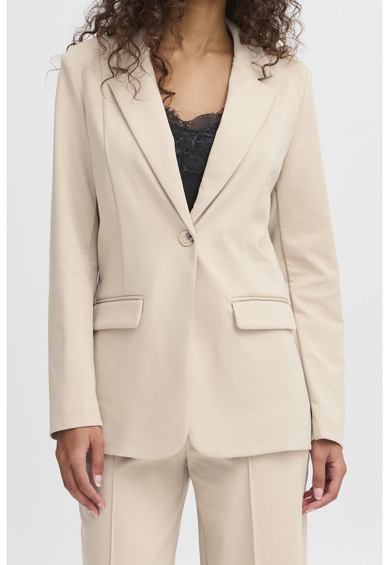Femme portant un blazer beige à un bouton et un pantalon assorti, avec un top en dentelle noire en dessous, debout devant un fond uni.