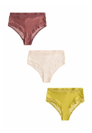 Žena s blond vlasmi po plecia nosi žlto-zelenú priehľadnú sieťovanú podprsenku a nohavičky s vysokým pásom, stojaca pred jednoduchou svetlou stenou.