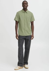 Camicia a maniche corte, verde chiaro, con bottoni e taschino sul petto, abbinata a jeans larghi grigio scuro e sneakers color crema.