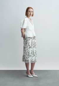 Λευκό κοντό τοπ με φουσκωτά μανίκια πάνω από πλισέ μίντι φούστα με σχέδιο λουλουδιών, συνοδευόμενο από λευκές μυτερές γόβες.