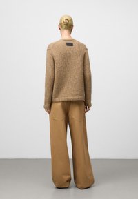 Brązowy sweter dziany z okrągłym dekoltem i opuszczonymi ramionami, noszony z luźnymi brązowymi spodniami z tylnymi kieszeniami.