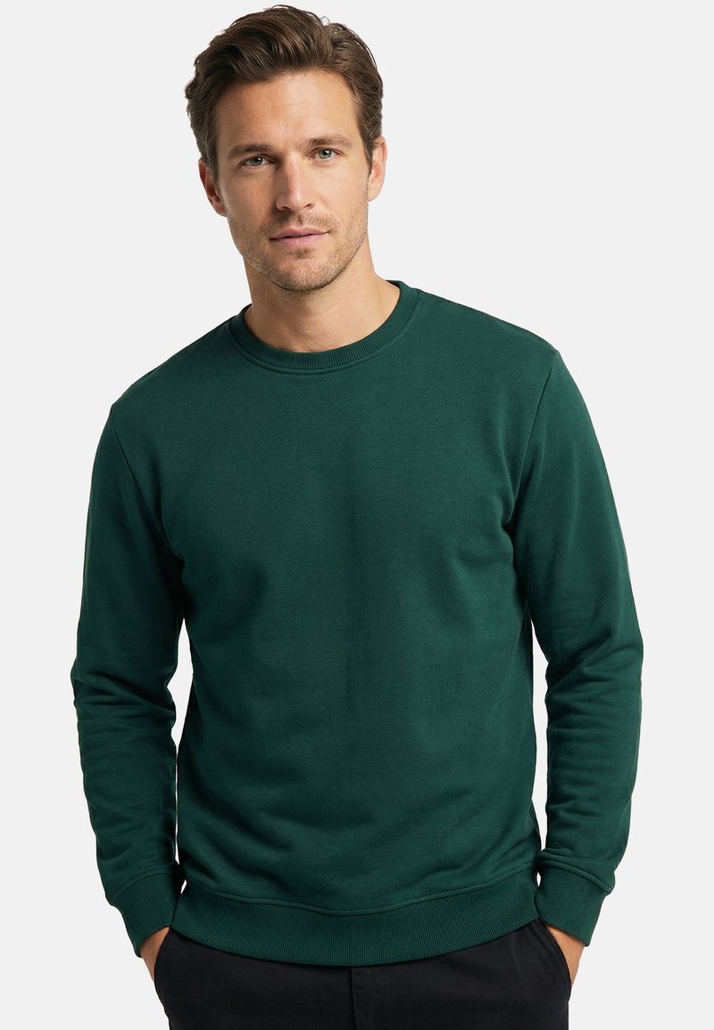 Homme aux cheveux bruns courts portant un sweat-shirt vert foncé à manches longues et col rond, les mains dans les poches, face à l'avant.