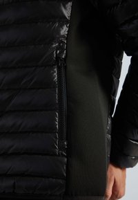 Veste matelassée noire avec panneaux texturés matelassés, tissu latéral contrastant vert et une poche zippée pour plus de fonctionnalité.