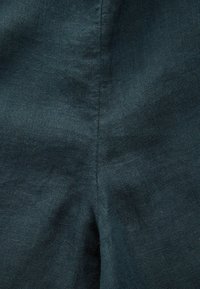 Ciemnozielona lniana tkanina, gładka w dotyku, z delikatnymi nieregularnościami i tkanym detalem. Zbliżenie pokazujące jakość naturalnego włókna.