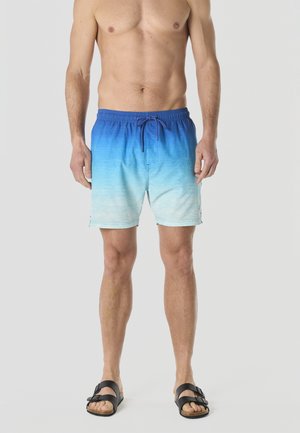 Hombre sin camiseta con pantalones de baño en degradado azul y sandalias negras con hebilla, de pie frente a un fondo liso.