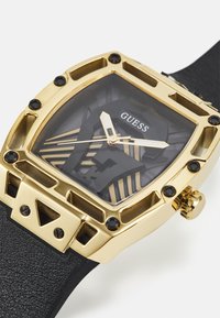 Kullatud ruudukujuline kell musta mustriga sihvriga, mineraalklaasiga ja tekstureeritud musta kummirihmaga. Omab logo ja musti aktsente.