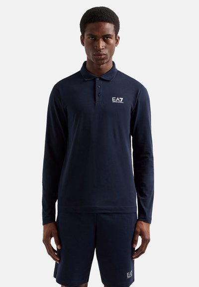 Jeune homme portant un polo manches longues bleu marine et un short assorti avec les logos EA7 Emporio Armani, debout sur fond blanc.