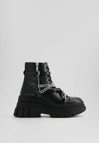 Botte de combat noire à semelle épaisse, avec fermeture éclair, sangles à boucle, décoration en chaîne argentée et semelle crantée épaisse sur un fond gris uni.