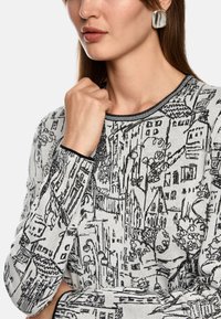 Knit sweater featuring a black and white cityscape pattern, round neckline, and contrasting black trim. Model wearing a large earring.