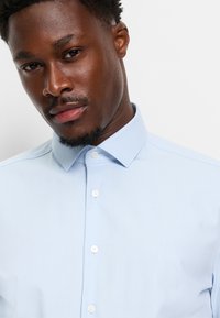 Camicia azzurra chiara con bottoni bianchi e colletto classico, dal tessuto liscio. Vista ravvicinata del collo e del colletto.