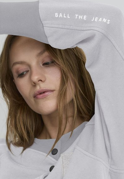 Light gray sweatshirt featuring ribbed cuffs and a textured pocket, with "BALL THE JEANS" printed in white on the sleeve.