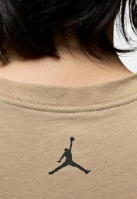 T-shirt color khaki con scollo rotondo e maniche corte. Presenta un logo Jumpman nero stampato sulla parte superiore della schiena. Texture in cotone morbido.