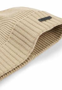 Cappello di lana beige lavorato a maglia con una texture a coste, caratterizzato da un piccolo etichetta nera. Il tessuto è morbido ed elastico, progettato per offrire calore e comfort.