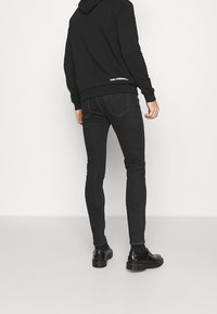 Jeans skinnies de mezclilla negra con una textura suave, costuras sutiles y dos bolsillos traseros, combinados con una sudadera negra y zapatos negros.