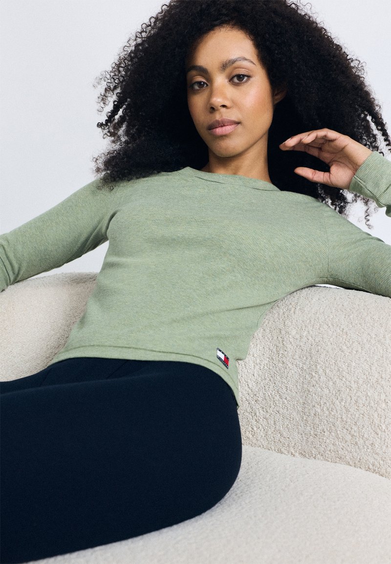 Olijfgroene, geribbelde top met lange mouwen en een ronde halslijn, voorzien van een klein logopatch op de linkeronderkant. Gepaard met donkere leggings.