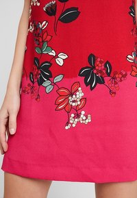 Rotes Blumenkleid mit einem Farbverlauf zu Pink am Saum. Verfügt über schwarz-weiße Blumenmuster mit grünen Blättern. Glattes, leichtes Material.