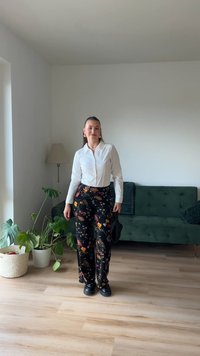 White button-up shirt paired with black floral-patterned wide-leg trousers. Black shoes, standing in a room with plants and a green sofa.
