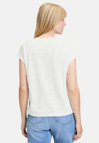 Hvid t-shirt med korte ærmer og et tekstureret mønster på ryggen, vist sammen med lyseblå jeans. Enkel, afslappet design.