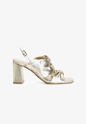 Kultainen metallinen korkokenkä, jossa on neliön muotoinen kärki, solmittu etusuunnittelu ja paksu korko. Mukana säädettävä nilkkaremmi.