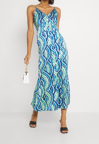 Robe maxi à motifs bleus et verts avec des bretelles spaghetti, un décolleté en V et des boutons ; associée à des talons ouverts noirs et un sac à main blanc.
