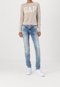 Pull en beige à côtes avec le logo "GAP" ; jeans déchirés bleu clair ; baskets blanches ; coupe décontractée, jambe droite, détails de couture visibles.