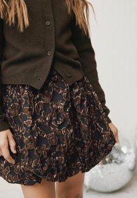 Cardigan lavorato a maglia marrone scuro con bottoni rotondi, abbinato a una gonna nera con un motivo floreale testurizzato in diverse tonalità di marrone e dettagli lucidi.