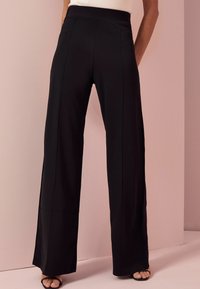 Black wide-leg pants with a smooth texture, high waist, and side seams, featuring a relaxed fit and minimal design, paired with black sandals.