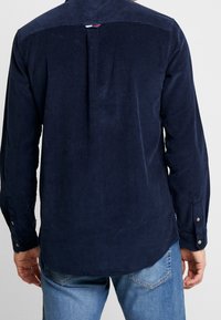 Marineblauwe corduroy blouse met lange mouwen, met knoopmanchetten en een achterplooi; heeft een contrasterende lus bij de halslijn voor extra detail.