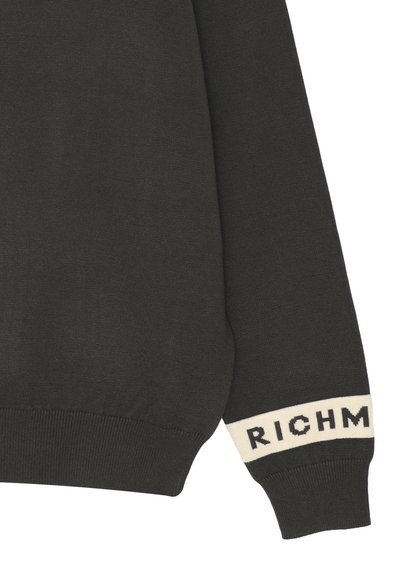 Manica di un maglione lavorato a maglia di un verde scuro, con una texture a coste, caratterizzata da un polsino crema a contrasto con la scritta "RICH" in grassetto nero.