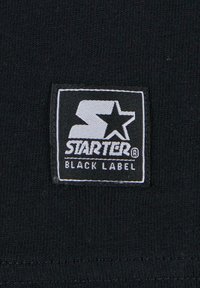 Juoda medžiaga su stačiakampiu baltu etikete, kurioje juodu šriftu parašyta "STARTER®" viršuje, o žvaigždė ir "BLACK LABEL" apačioje, tvarkingai siuvinėta.
