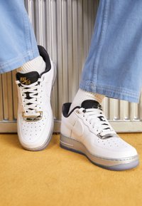 Sneakers Nike Air bianche con accenti argento, dettagli neri sulla linguetta e punta traforata, abbinate a pantaloni azzurri e calzini bianchi.